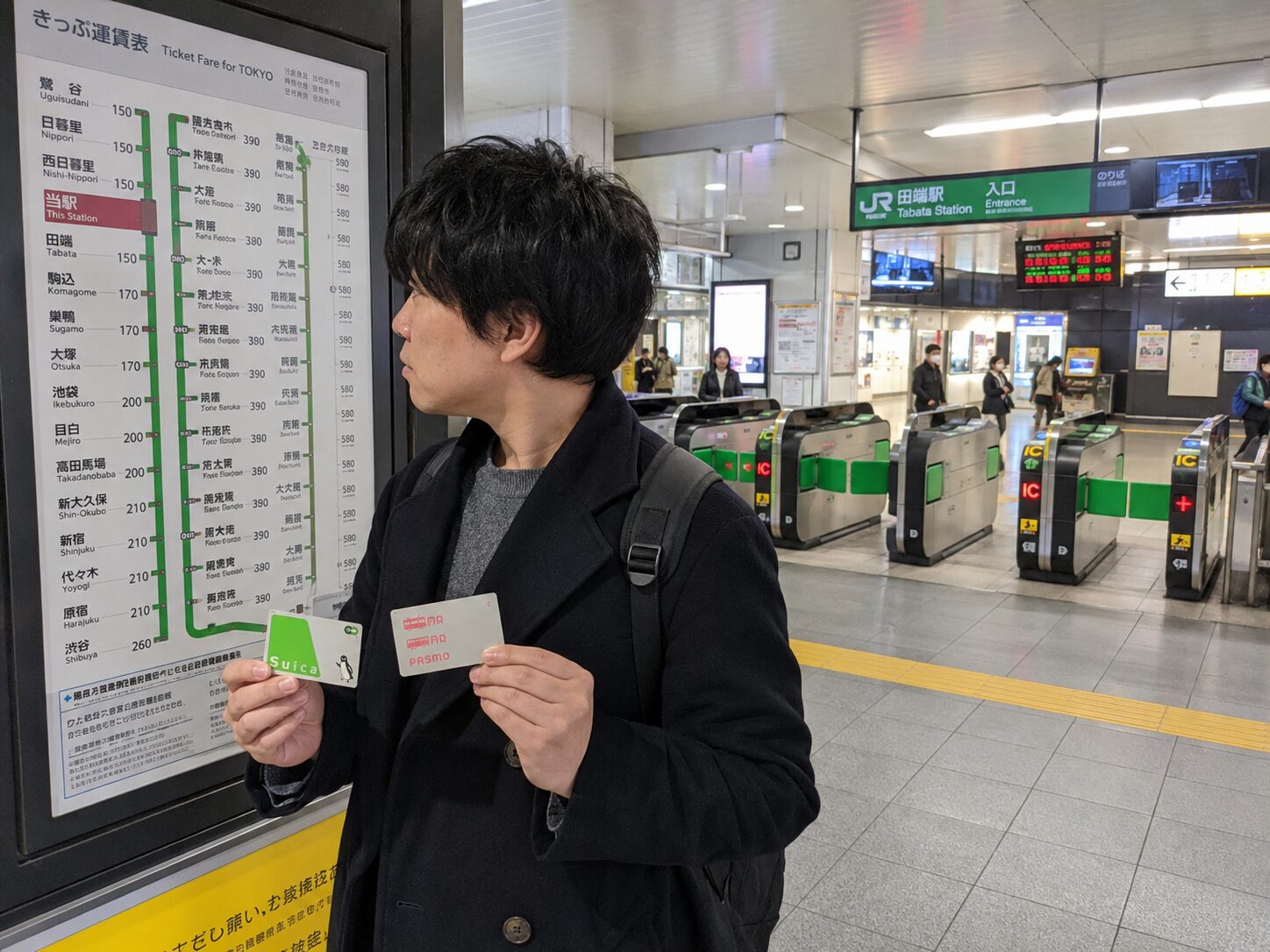 SuicaとPASMOを持った利用者が駅の料金案内と自動改札を確認している写真