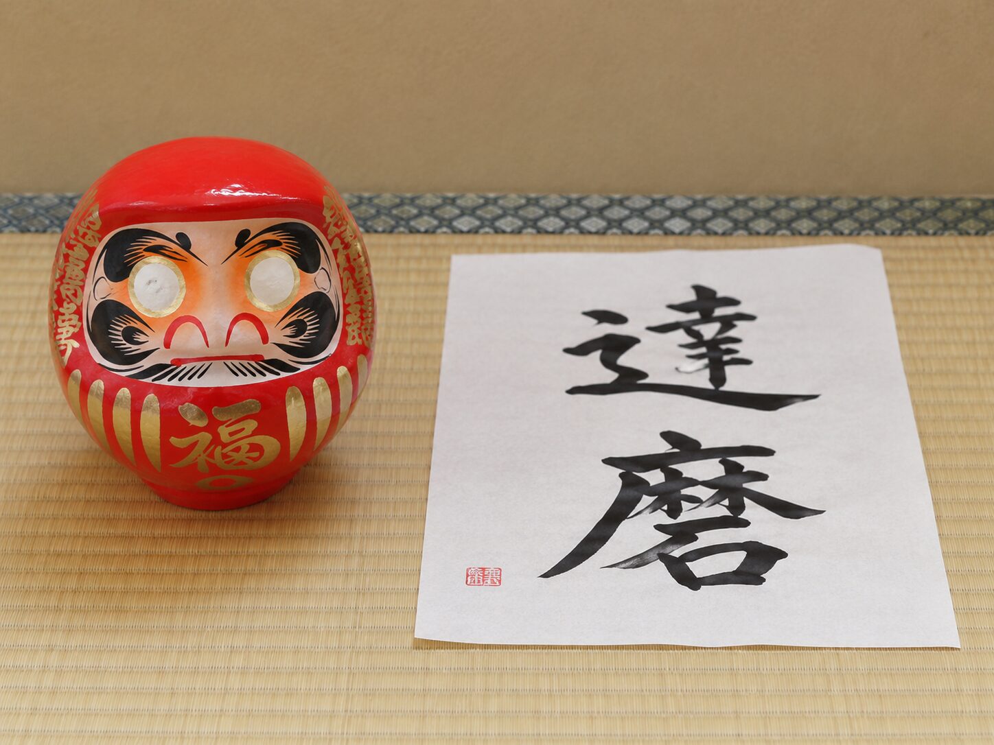 赤いだるま人形の横に筆文字で達磨と書かれた和紙が置かれている写真