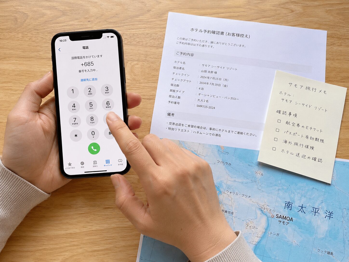 スマートフォンのダイヤル画面に+685から始まる番号を入力し、横にサモア旅行の予約確認書、ホテル名のメモ、南太平洋の地図が置かれているイメージ。無料写真素材で、手元、スマホ、旅行書類、国際電話の雰囲気が分かる画像を使用する