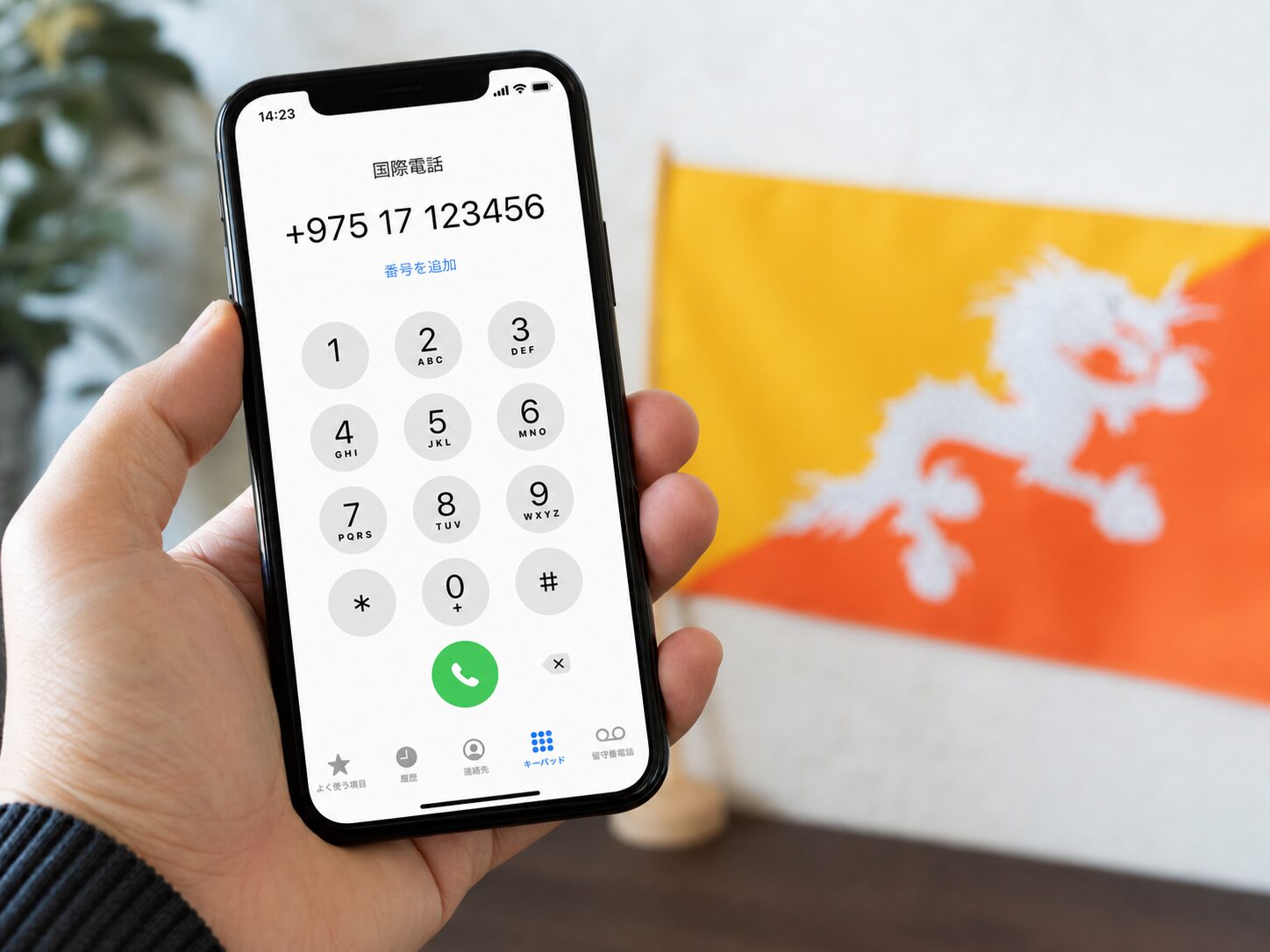 日本からブータンへ国際電話をかける場面。スマートフォン画面に+975とブータンの電話番号が表示され、背景にブータン国旗がある写真