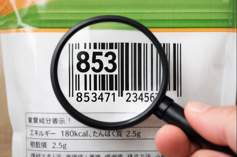 商品パッケージのバーコード番号を拡大して先頭3桁(85x)を確認しているイメージ画像(無料素材の想定)