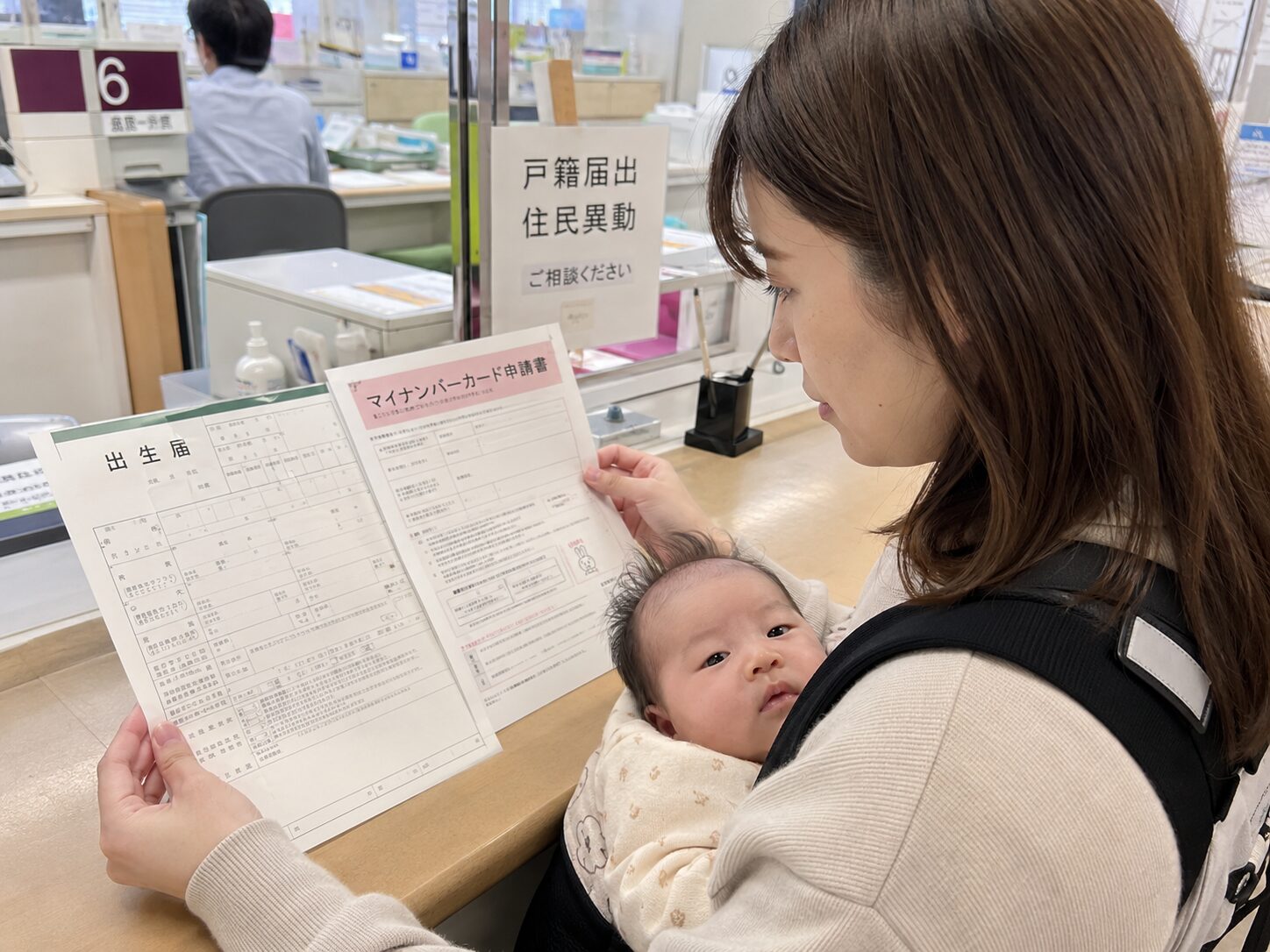 赤ちゃんを抱いた保護者が市区町村の窓口で出生届とマイナンバーカード申請書を確認している写真